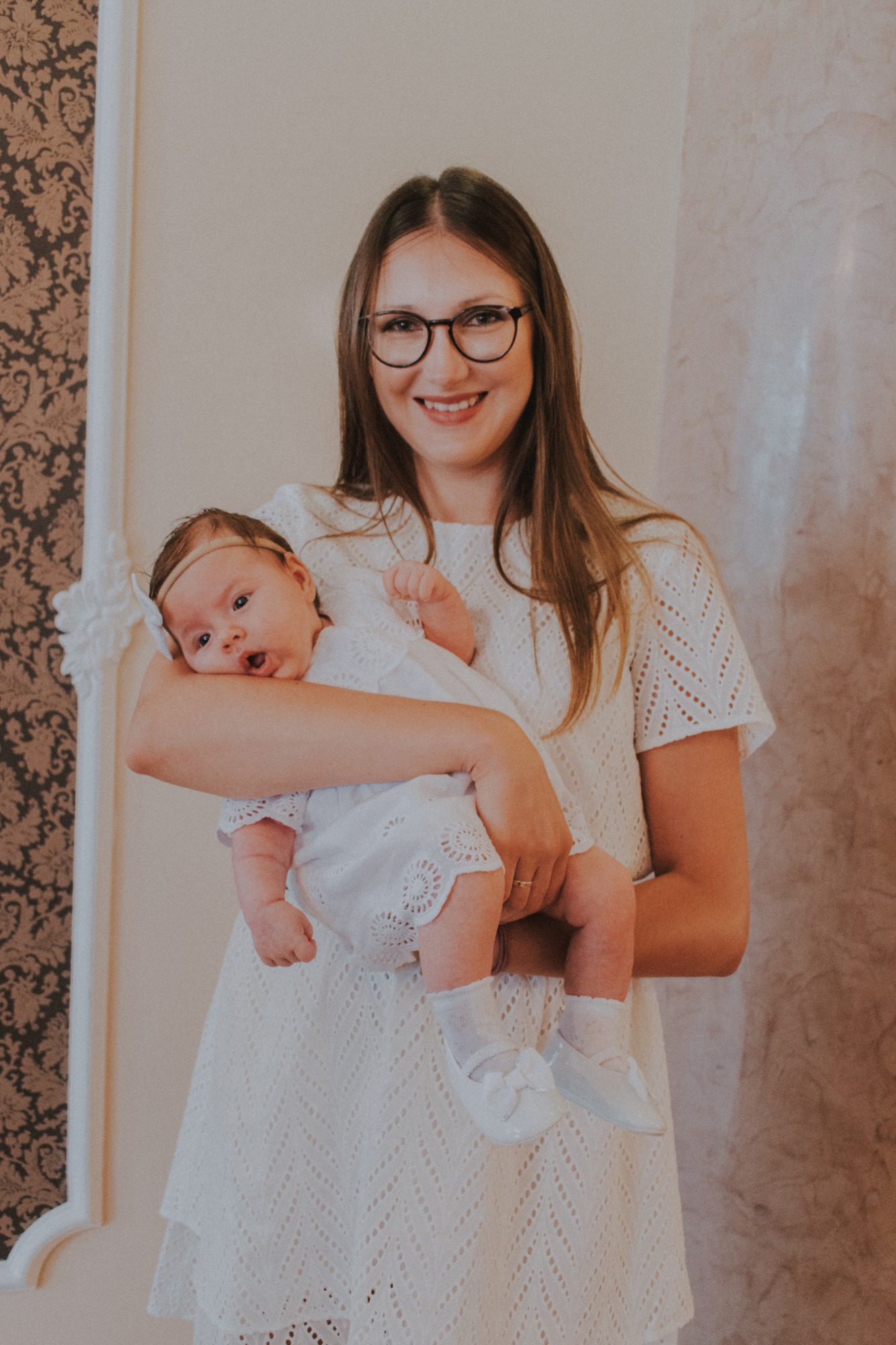 Uśmiechnięta kobieta w okularach trzyma na rękach niemowlę w białej sukience i bucikach z kokardkami, na tle ściany z dekoracyjnym wzorem.