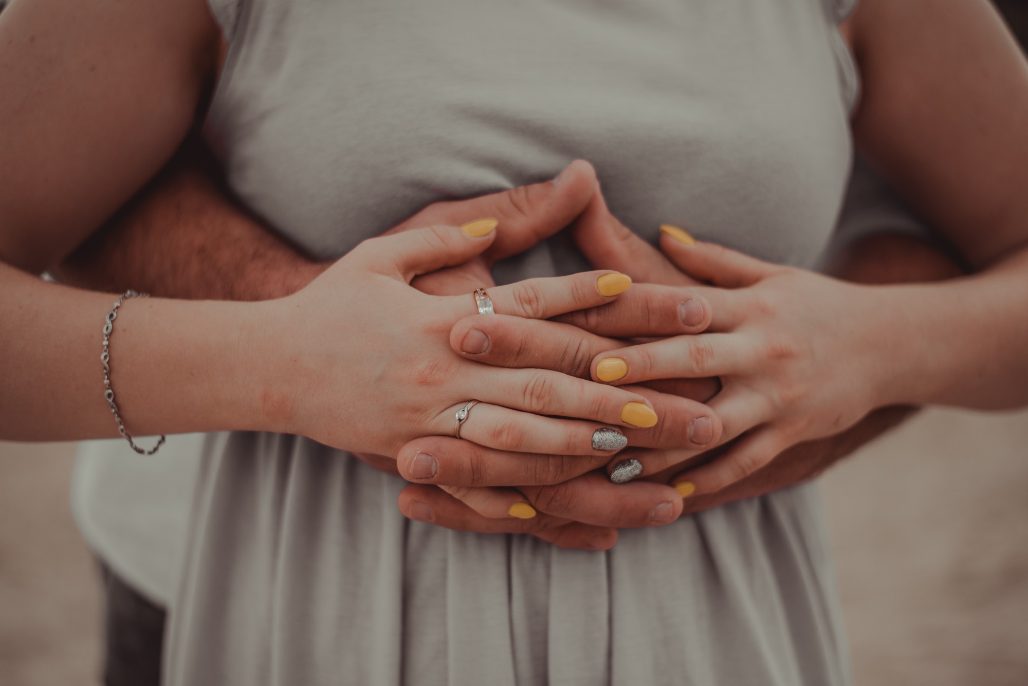 Zbliżenie dłoni pary: dłoń kobiety z żółtym manicure i srebrnym brokatowym akcentem na jednym paznokciu, oraz pierścionkiem, spoczywa na dłoni mężczyzny, obejmującej ją. W tle widoczny fragment...