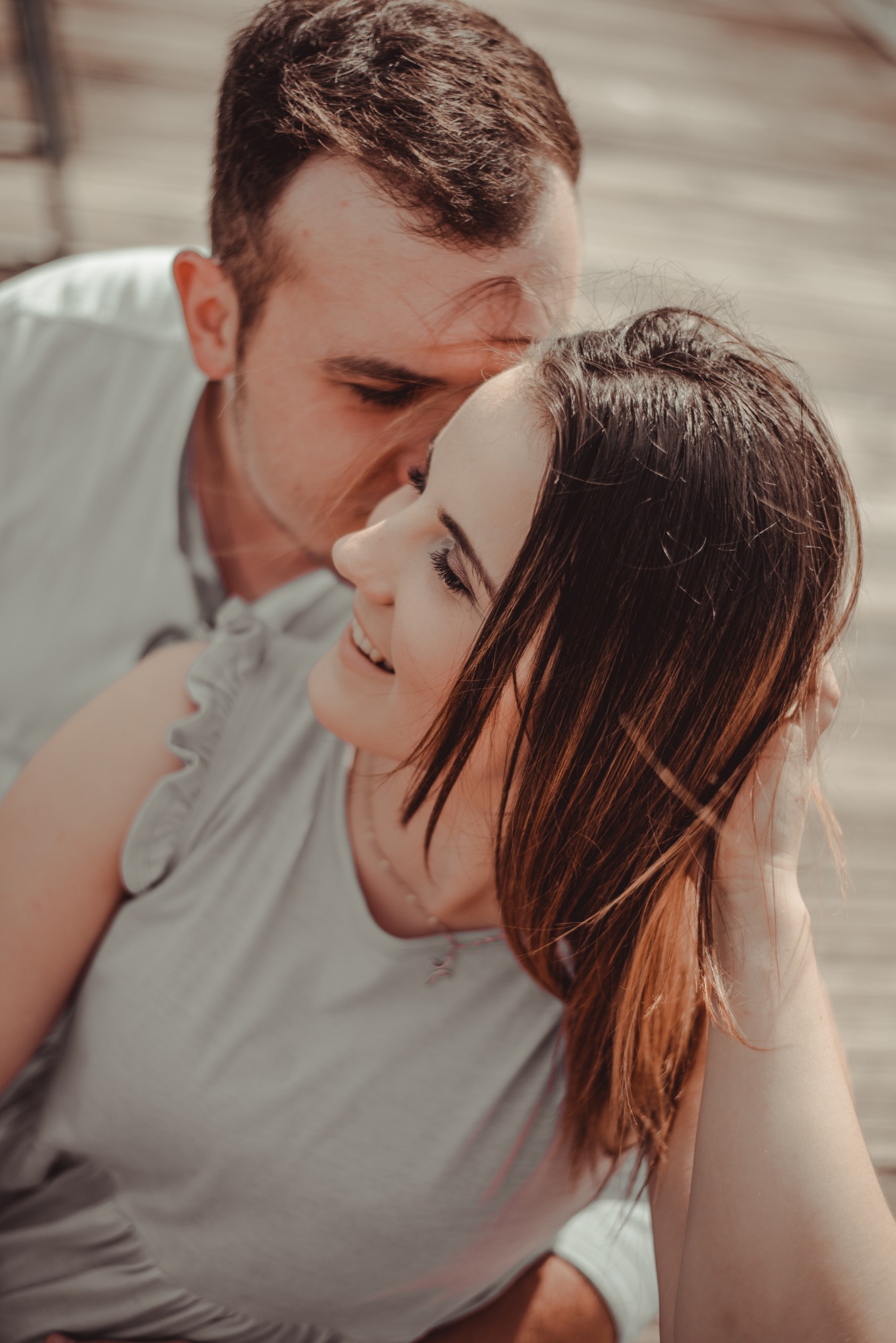 Uśmiechnięta kobieta z brązowymi włosami, mężczyzna całuje ją w skroń na tle drewnianej podłogi.