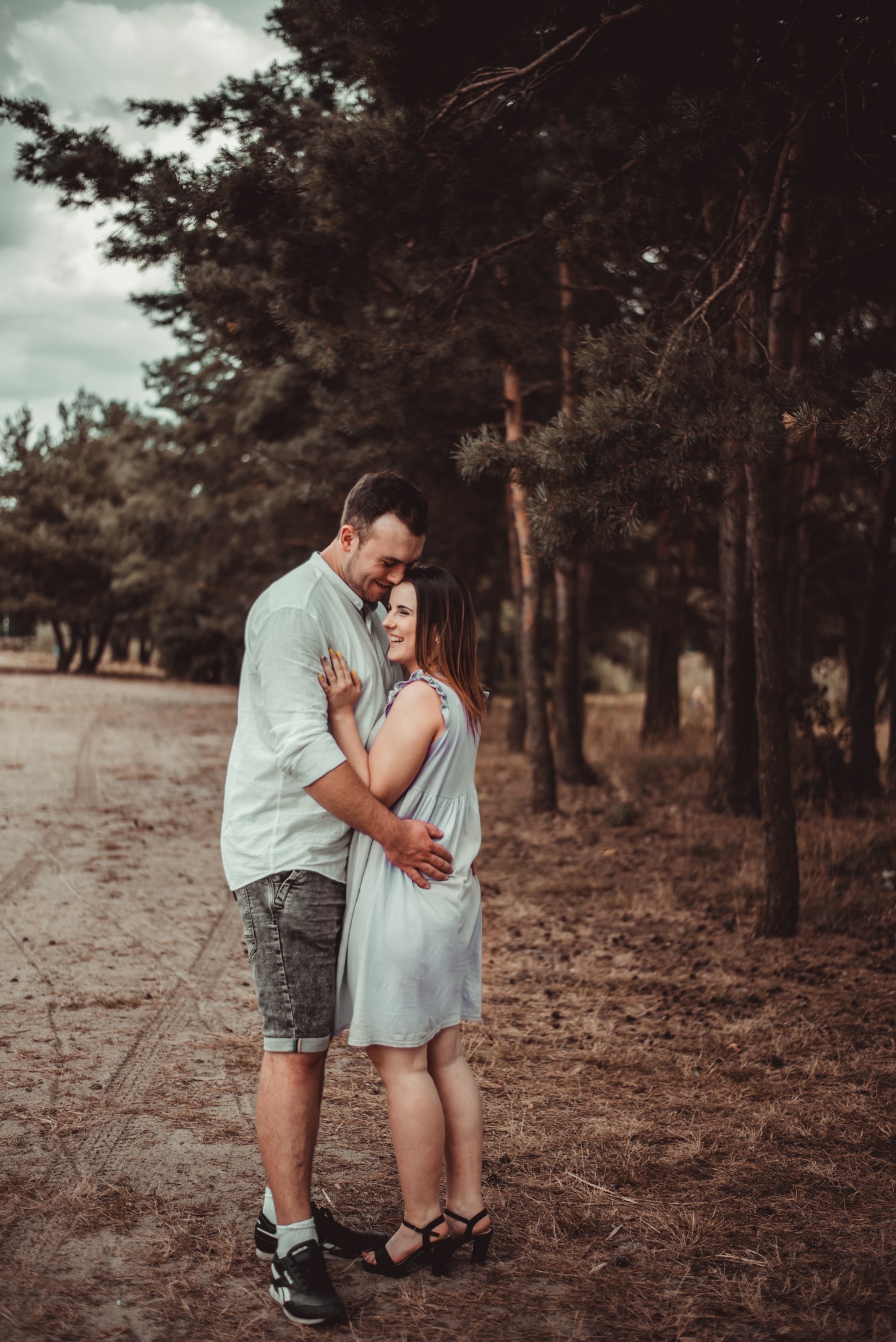 Uśmiechnięta para w objęciach na tle leśnej ścieżki, on w białej koszuli i jeansowych szortach, ona w błękitnej sukience z falbankami, delikatne światło.