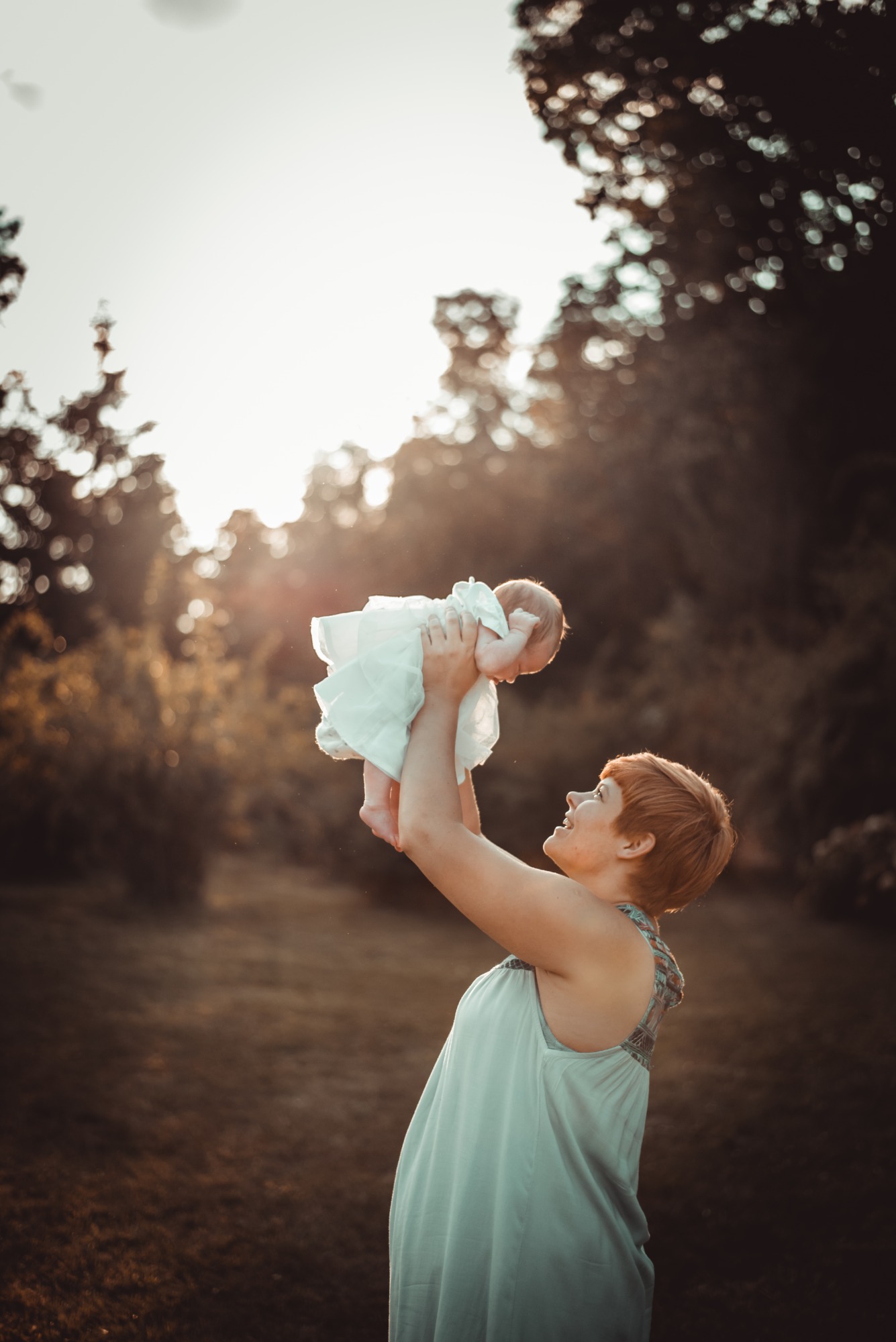 Mama z krótkimi rudymi włosami podnosi w górę niemowlę ubrane w białą sukienkę na tle drzew i zachodzącego słońca.