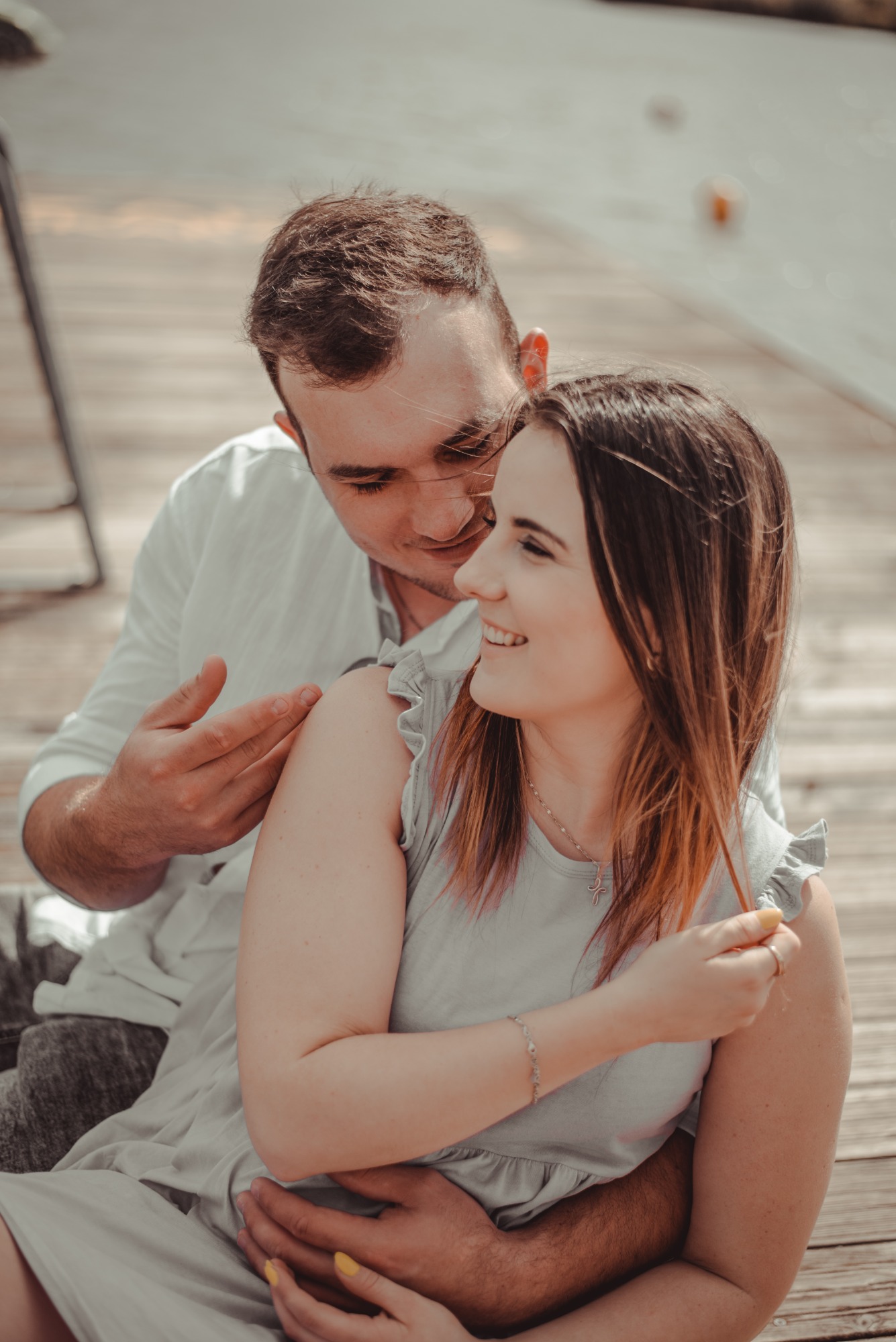 Uśmiechnięta para siedzi na drewnianym pomoście, mężczyzna obejmuje kobietę, która bawi się kosmykiem włosów.