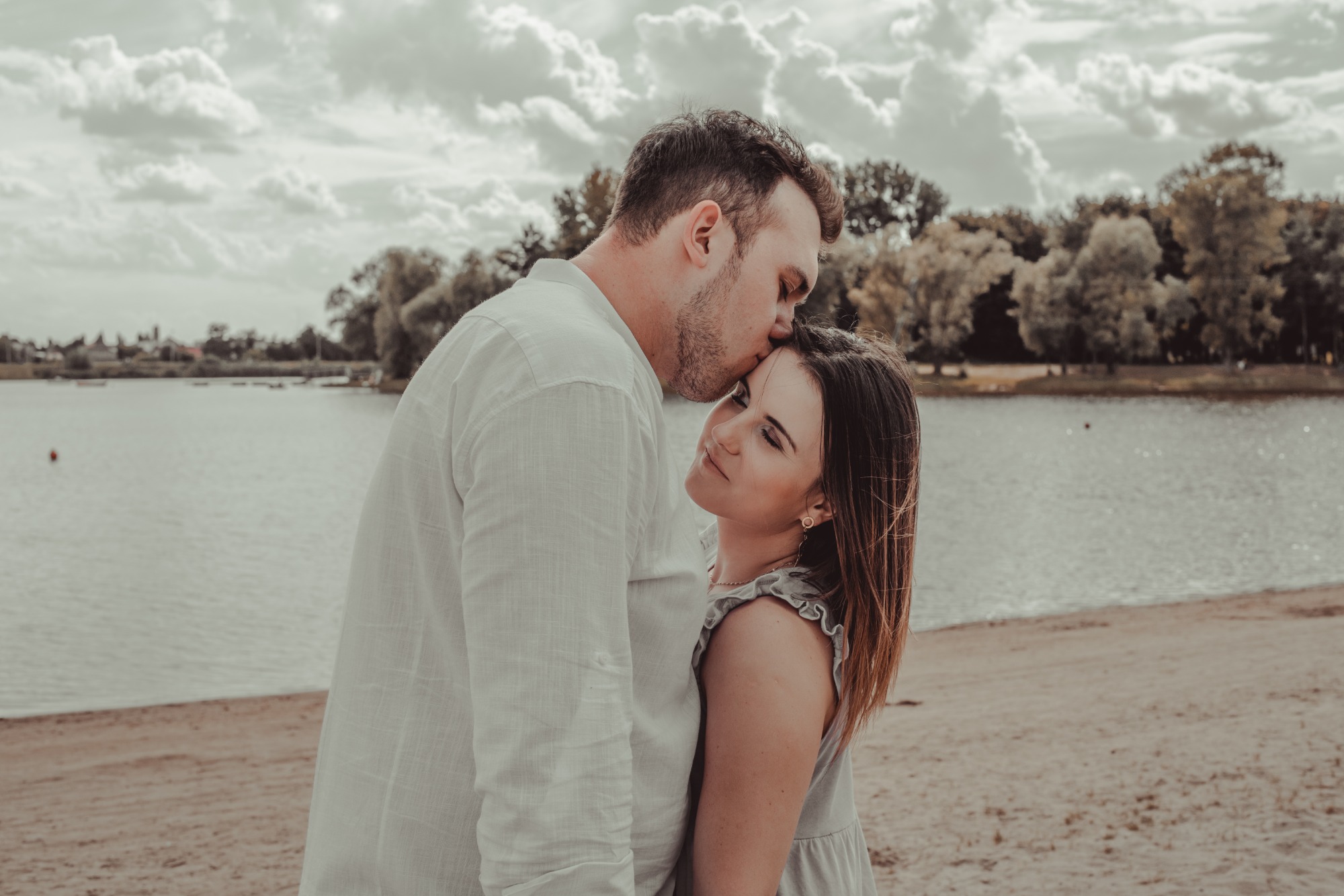 Mężczyzna w białej koszuli całuje kobietę w czoło na tle jeziora i pochmurnego nieba.