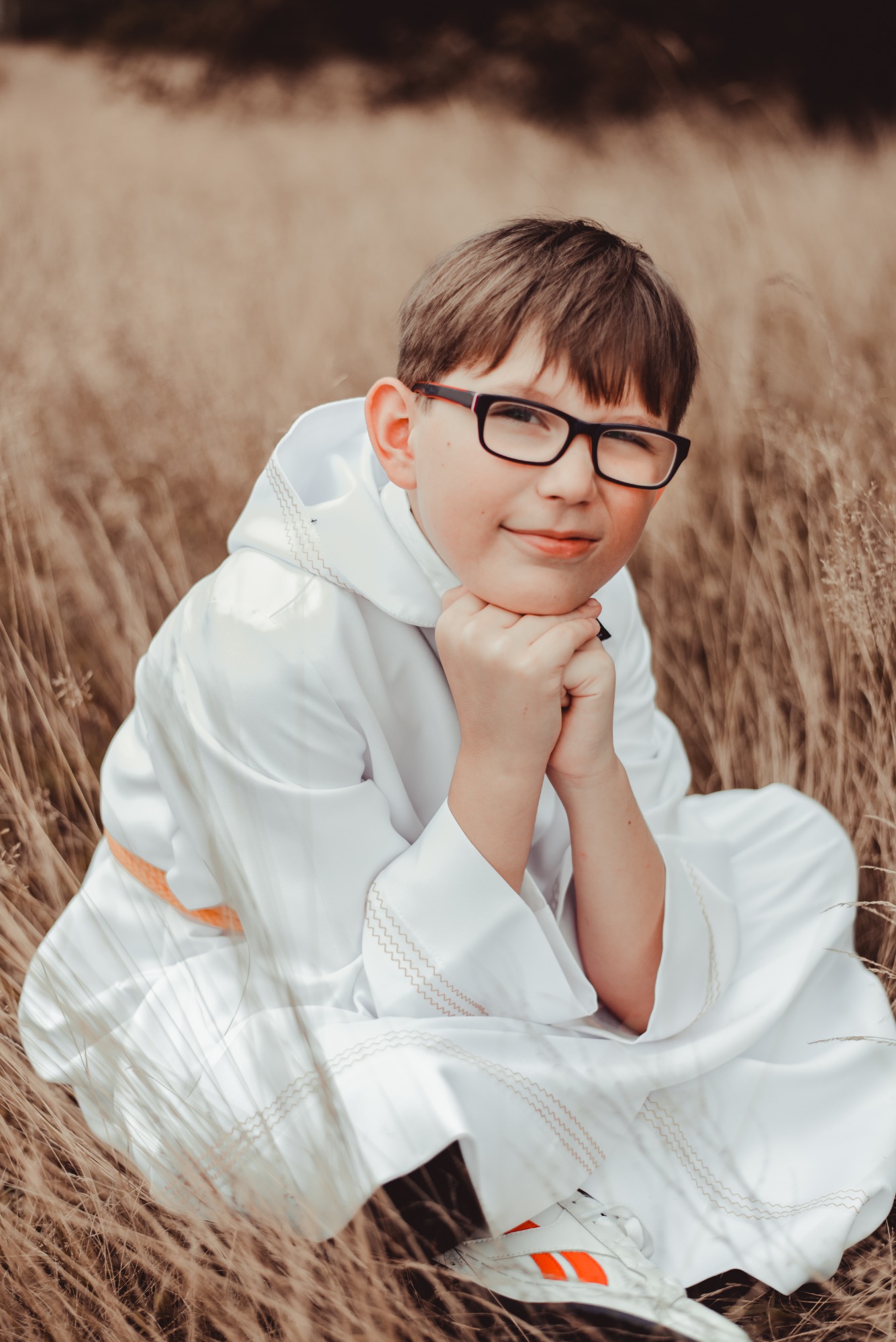 Chłopiec w stroju komunijnym, siedzący w trawie, z rękami pod brodą i okularami na nosie.
