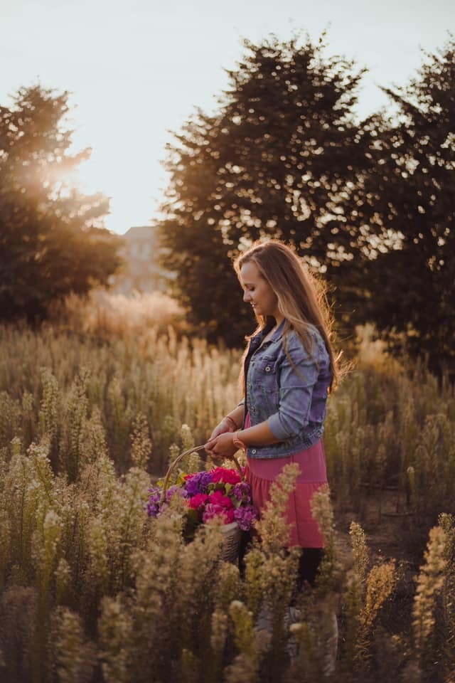Młoda kobieta z długimi włosami w dżinsowej kurtce i różowej sukience niesie koszyk pełen różowych i fioletowych kwiatów na łące w promieniach zachodzącego słońca.