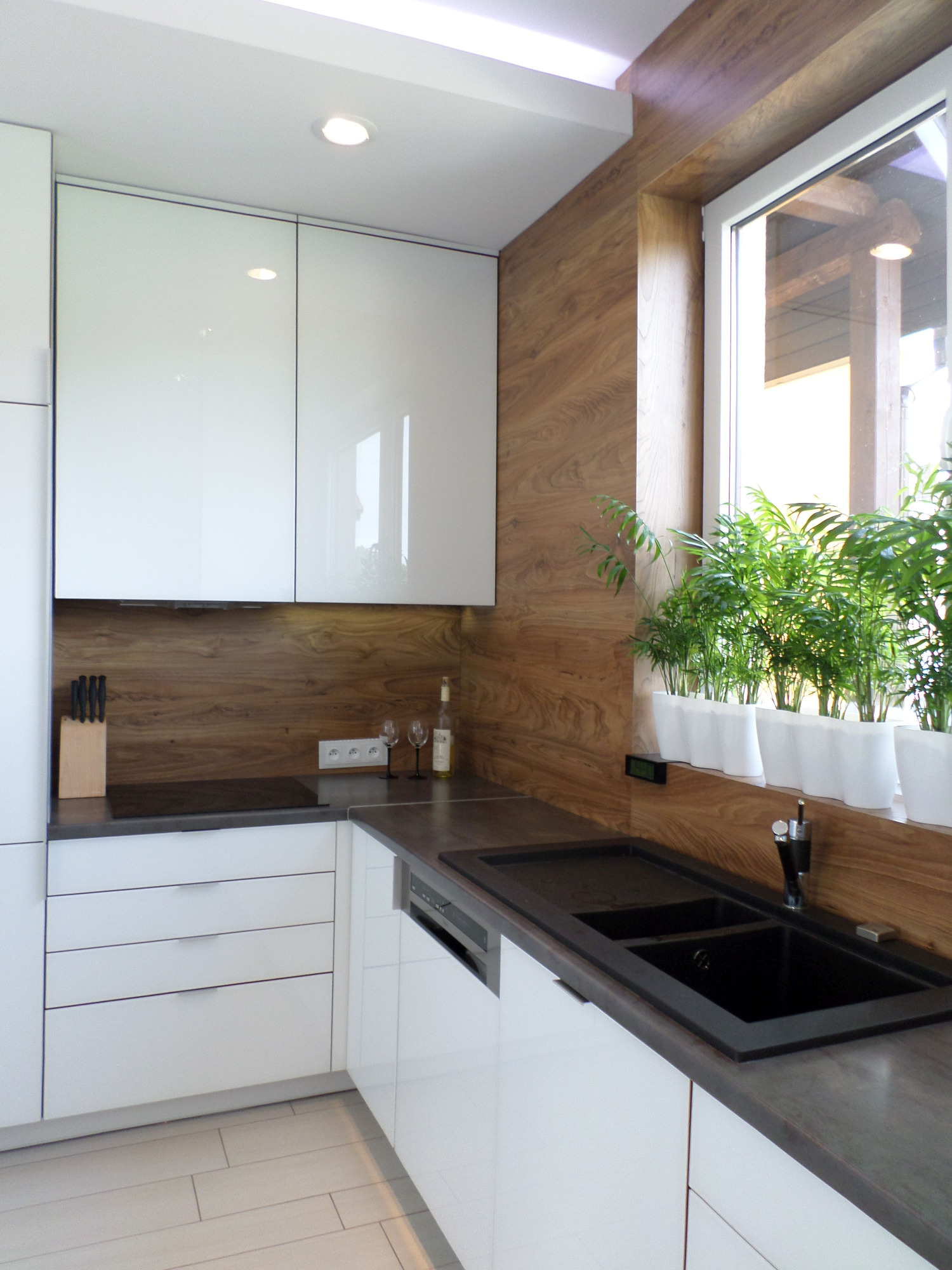 Nowoczesna kuchnia z białymi, lakierowanymi szafkami, blatem w kolorze grafitu, drewnianą okładziną ściany i oknem z roślinami doniczkowymi.