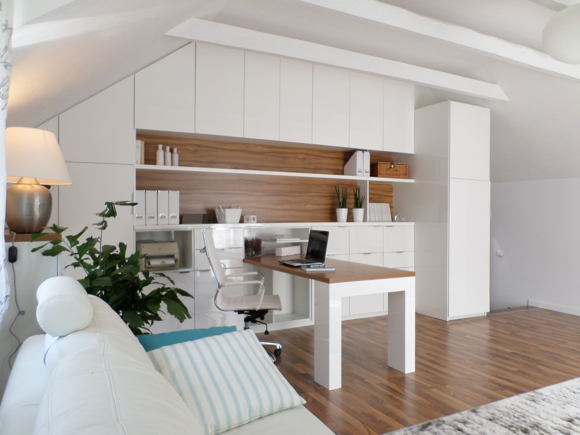 Jasny gabinet na poddaszu z biurkiem, laptopem, białymi meblami w zabudowie i drewnianymi akcentami, widok z perspektywy sofy z poduszkami.