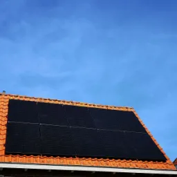 Panele słoneczne zamontowane na pomarańczowym dachu z dachówki ceramicznej, widok z dołu na tle błękitnego nieba.