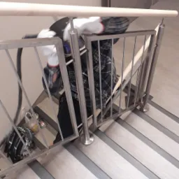 Montaż balustrady nierdzewnej z drewnianą poręczą na schodach, widok z góry, pracownik w koszuli w kratę i rękawicach ochronnych, w tle drzwi.