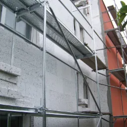 Budynek w Strzelinie w trakcie ocieplania, widoczne rusztowania i warstwa izolacyjna z szarego styropianu z czarnymi plamkami. Fragment ściany pomalowany na pomarańczowo.