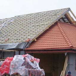 Protech - Budowa dachu domu jednorodzinnego: widoczne drewniane elementy konstrukcyjne, częściowo pokryte czerwoną dachówką, z zapasem dachówek na palecie w folii ochronnej.