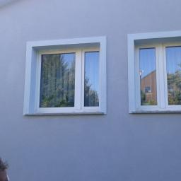 Protech - Jasnoszara elewacja z dwoma białymi oknami z jasnoniebieskimi ramami, w których odbijają się drzewa i budynek.