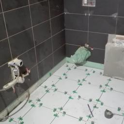 Protech - Wnętrze łazienki w trakcie prac wykończeniowych, widoczne czarne płytki ścienne i białe podłogowe z zielonymi klipsami poziomującymi, narzędzia glazurnicze na półce.