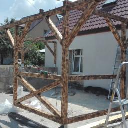 Protech - Konstrukcja drewniana z belek opalanych ogniem, tworząca zadaszenie tarasu, w trakcie montażu przy białej ścianie domu z brązową dachówką. Widoczna drabina aluminiowa, narzędzia i materiały budowlane.