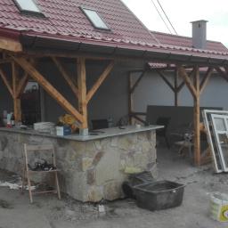 Protech - Zadaszona altana z kamienną podstawą baru w trakcie budowy, widoczne materiały budowlane i okna do montażu, dach z czerwonej dachówki.