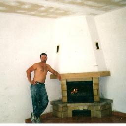 Protech - Mężczyzna bez koszulki w ubrudzonych farbą spodniach opiera się o kamienny kominek w rogu pokoju z niedokończonymi ścianami i sufitem.