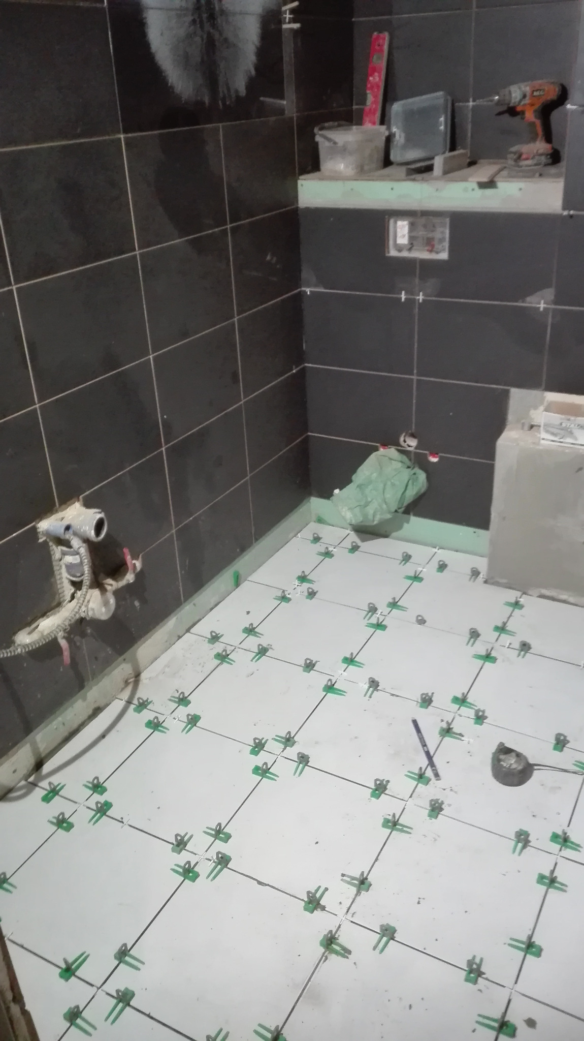 Wnętrze łazienki w trakcie prac wykończeniowych, widoczne czarne płytki ścienne i białe podłogowe z zielonymi klipsami poziomującymi, narzędzia glazurnicze na półce.