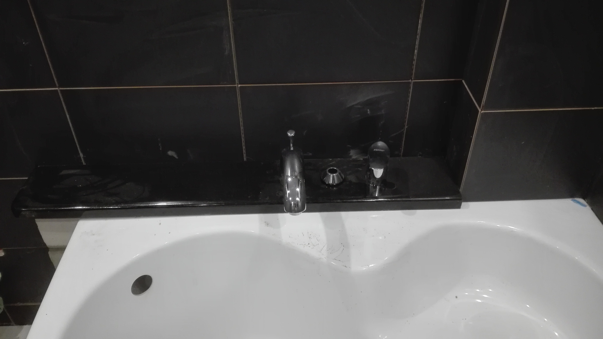 Biała umywalka z chromowaną baterią i czarną, błyszczącą płytką ceramiczną na ścianie, widoczne ślady po montażu.