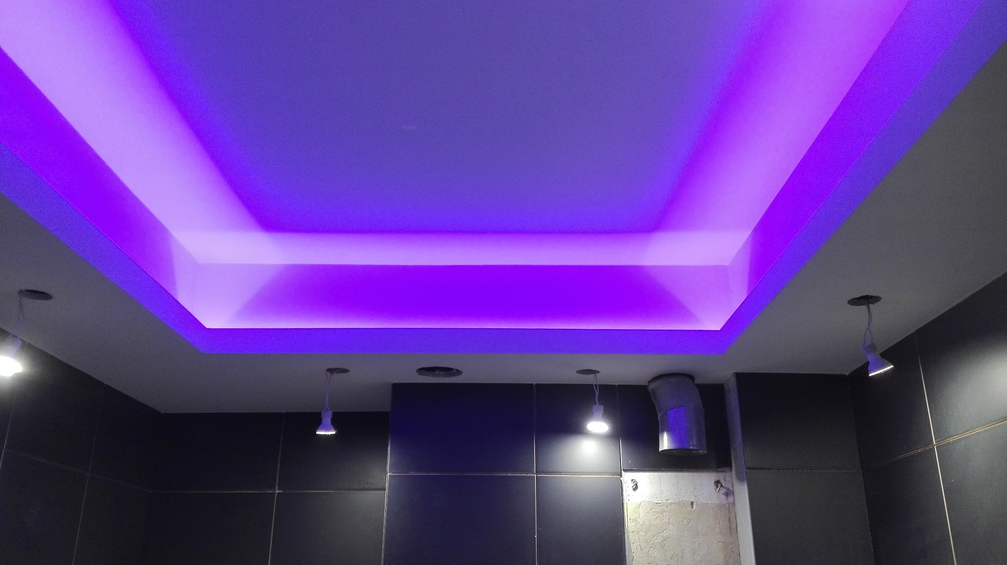Podwieszany sufit z dekoracyjnym oświetleniem LED w odcieniach fioletu, widoczne czarne płytki ścienne i wiszące lampy.