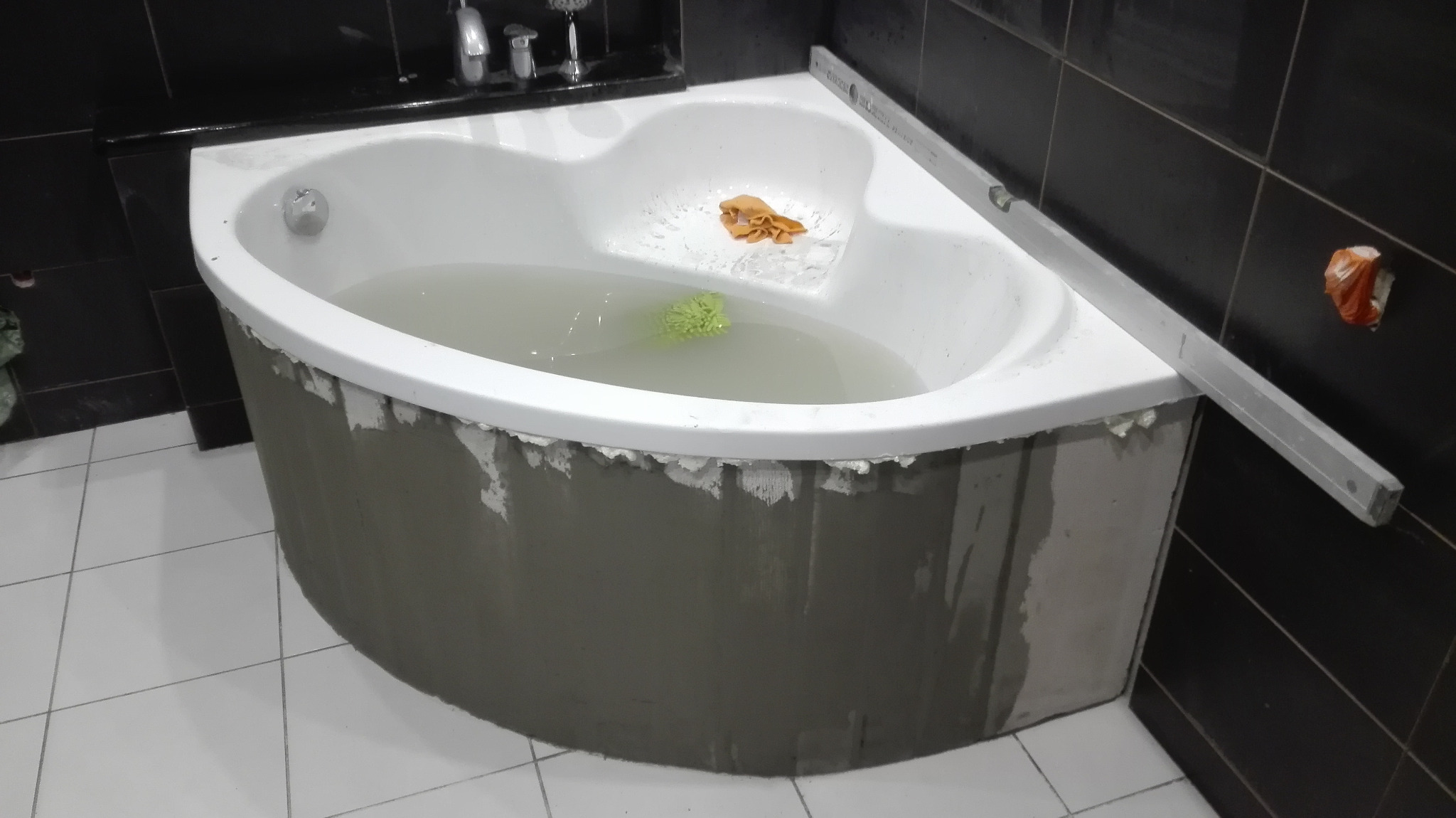 Wanna narożna wypełniona wodą, w trakcie obudowy, widoczny poziomica, czarne płytki na ścianie i białe na podłodze, wnętrze łazienki w trakcie prac wykończeniowych.