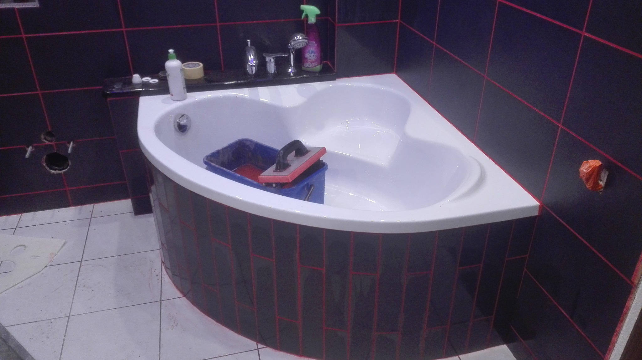 Narożna wanna obudowana czarnymi płytkami z czerwoną fugą, wiadro z narzędziami wewnątrz, czarne płytki na ścianach z czerwoną fugą, widoczne otwory w ścianie.