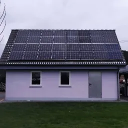 Dom z panelami fotowoltaicznymi na dachu, widok z przodu na trawnik i szarą fasadę, pochmurne niebo.