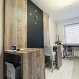 Nowoczesna kuchnia z drewnianym blatem barowym, czarną ścianą z zegarem i białymi krzesłami barowymi, oświetlona naturalnym światłem wpadającym przez okno z żaluzjami.