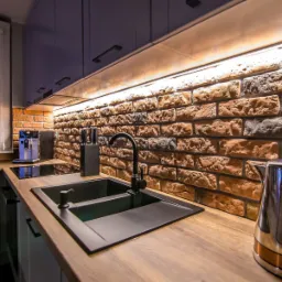 Nowoczesna kuchnia z czarnym, dwukomorowym zlewem, baterią i dozownikiem, drewnianym blatem, ścianą z cegły i oświetleniem LED podszafkowym.