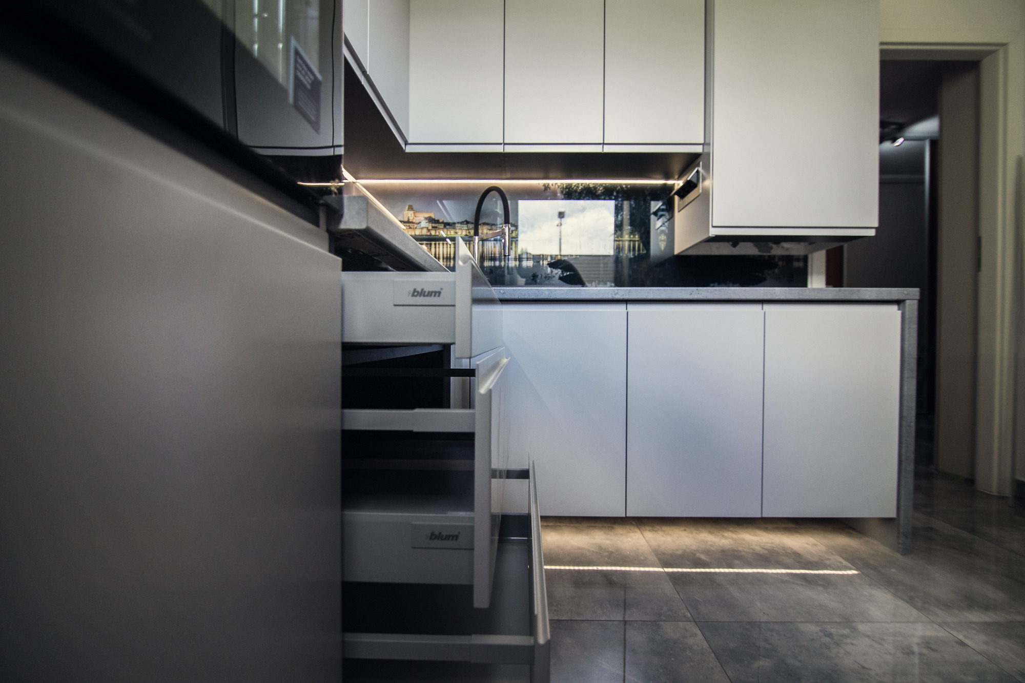 Nowoczesna zabudowa kuchenna z szufladami Blum, widok z otwartą szufladą, podświetlenie LED i dekoracyjny panel ścienny z motywem miasta w Toruniu.