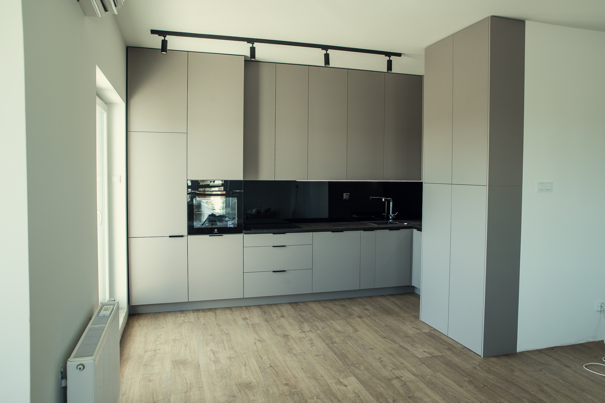 Nowoczesna kuchnia w odcieniach szarości z drewnopodobną podłogą, oświetleniem szynowym i zabudową sięgającą sufitu.