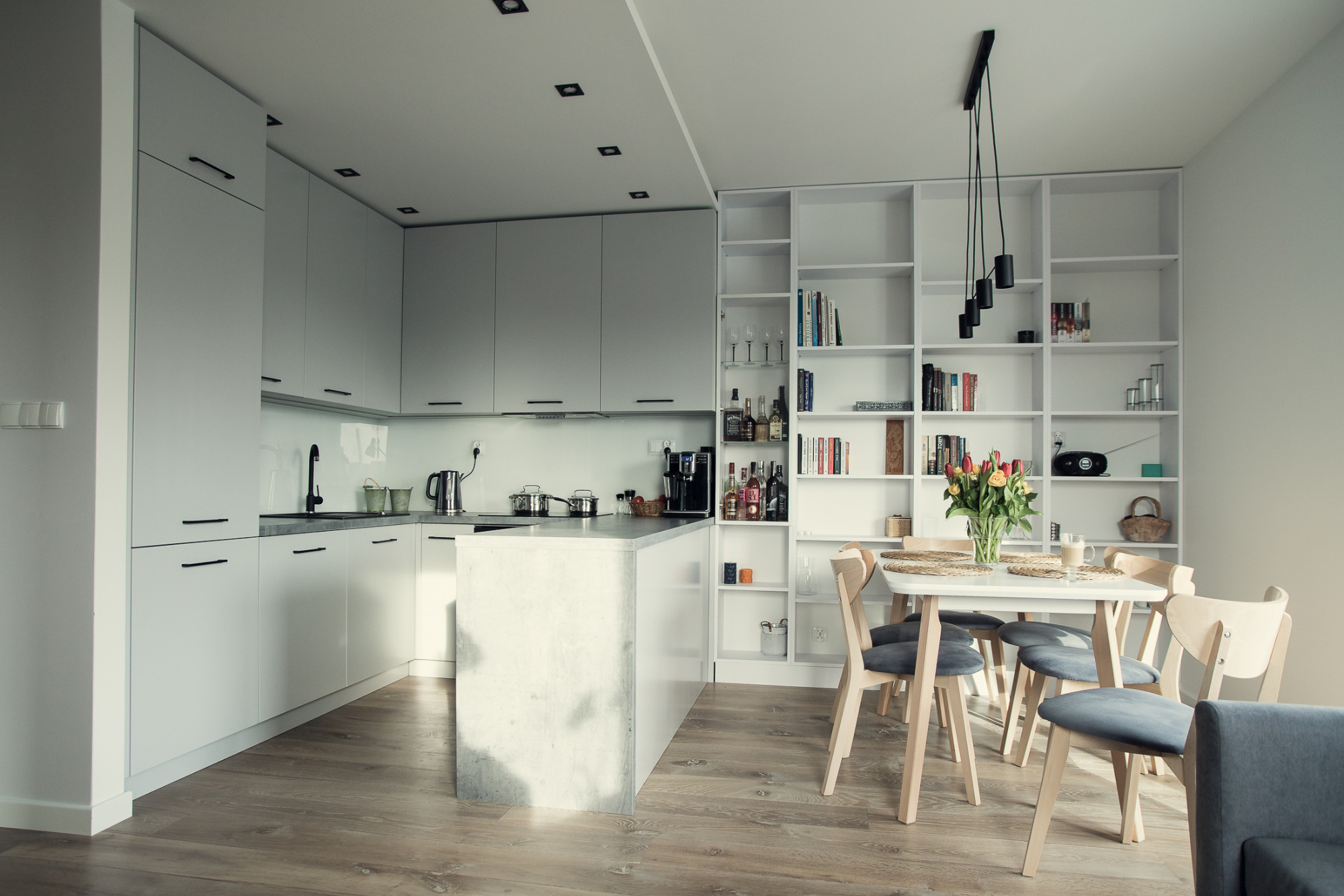 Jasna kuchnia z wyspą, białymi szafkami z czarnymi uchwytami, drewnianą podłogą i regałem na książki w tle, stół z bukietem tulipanów.