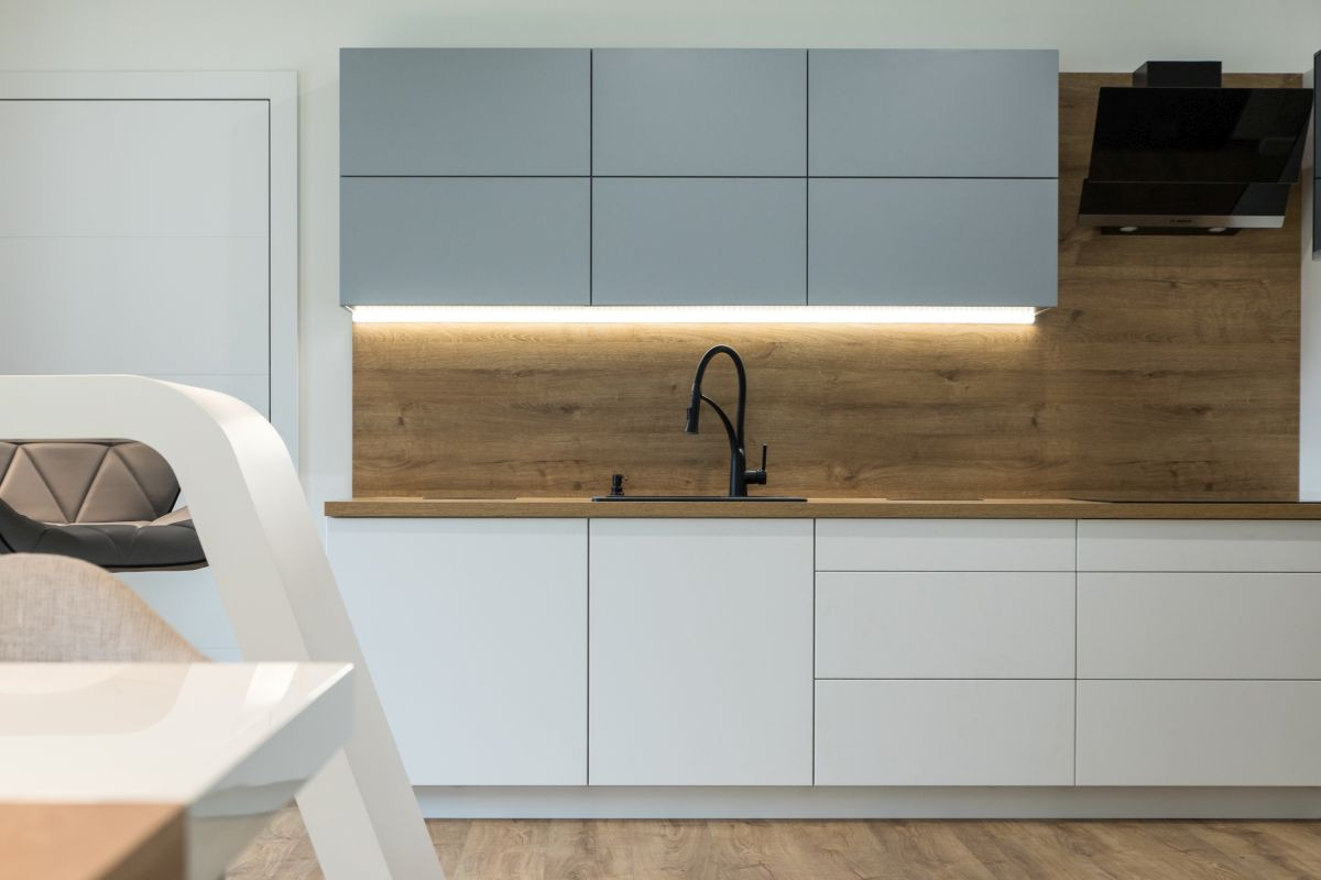 Nowoczesna kuchnia z białymi szafkami dolnymi i szarymi górnymi, drewniany panel ścienny z podświetleniem LED i czarną armaturą, fragment designerskiego krzesła w tle.