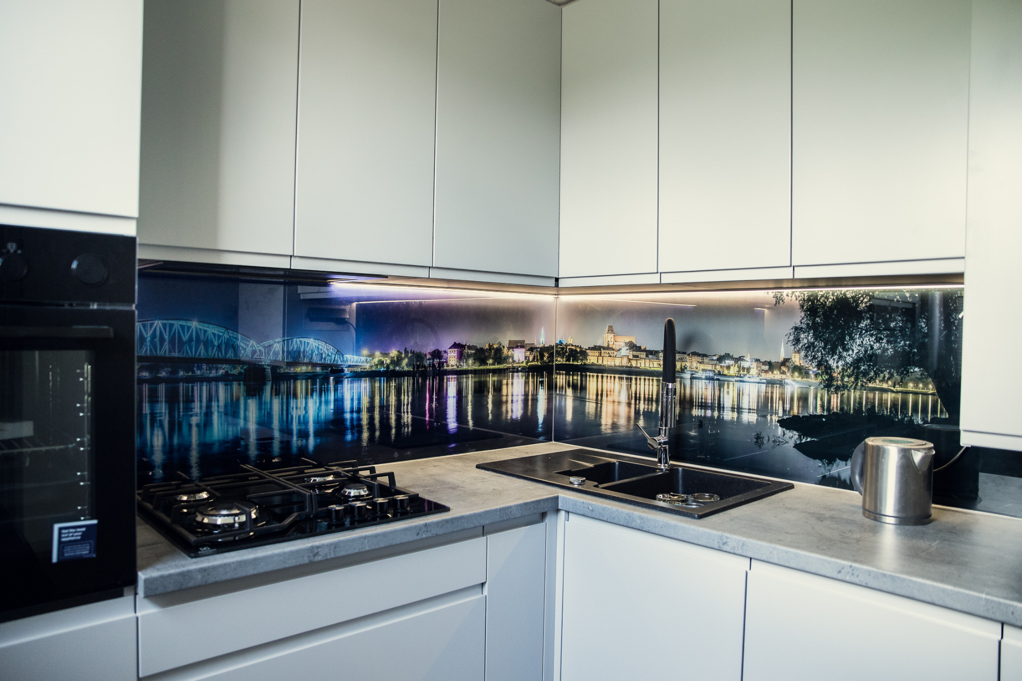 Nowoczesna kuchnia z białymi szafkami i blatem w odcieniach szarości, z czarnym zlewem i płytą gazową. Na ścianie nad blatem fototapeta z nocnym widokiem miasta odbijającego się w wodzie.
