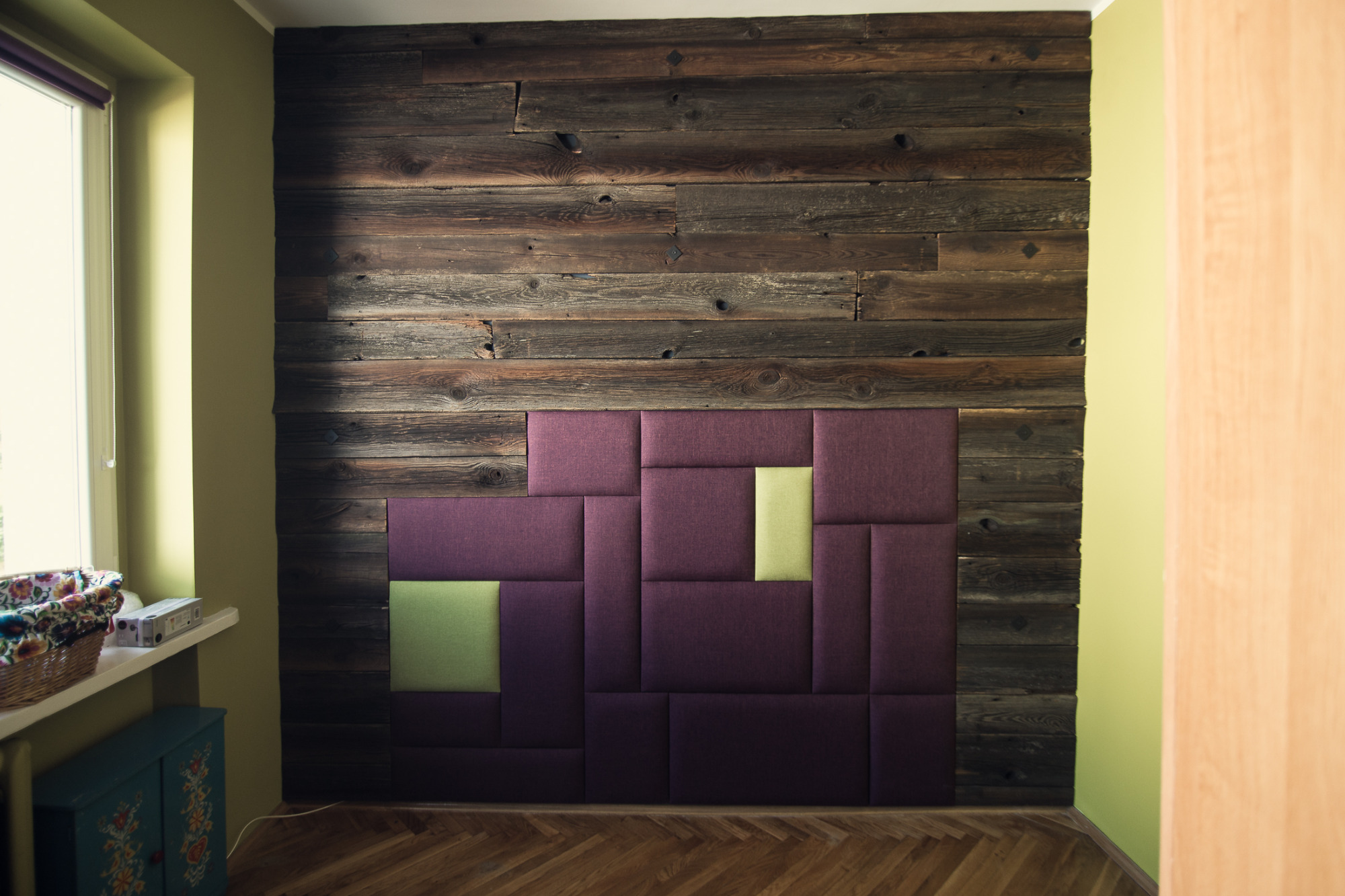 Tapicerowany panel ścienny w odcieniach fioletu i zieleni, ułożony z geometrycznych kształtów na tle ściany z ciemnego, postarzanego drewna.