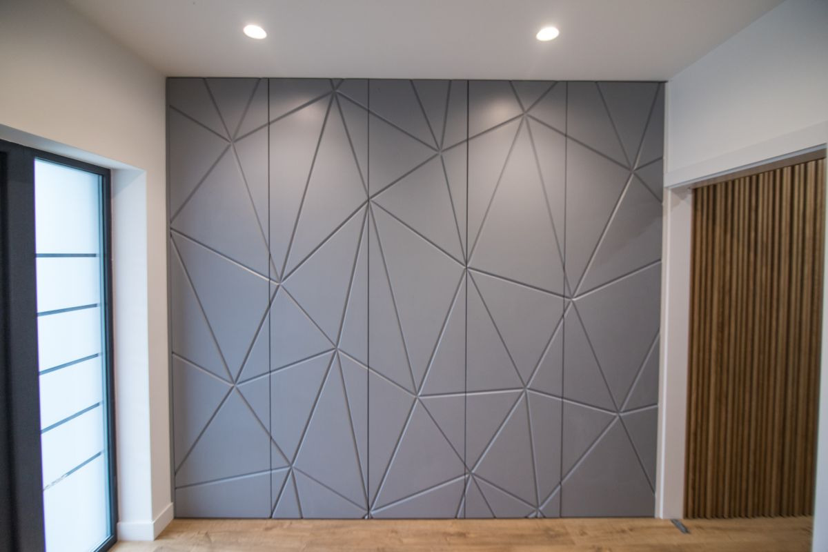Szara ściana z geometrycznym wzorem 3D, podświetlona punktowymi lampami sufitowymi, widok na drewnianą podłogę i fragmenty okna oraz drzwi z lamelami.