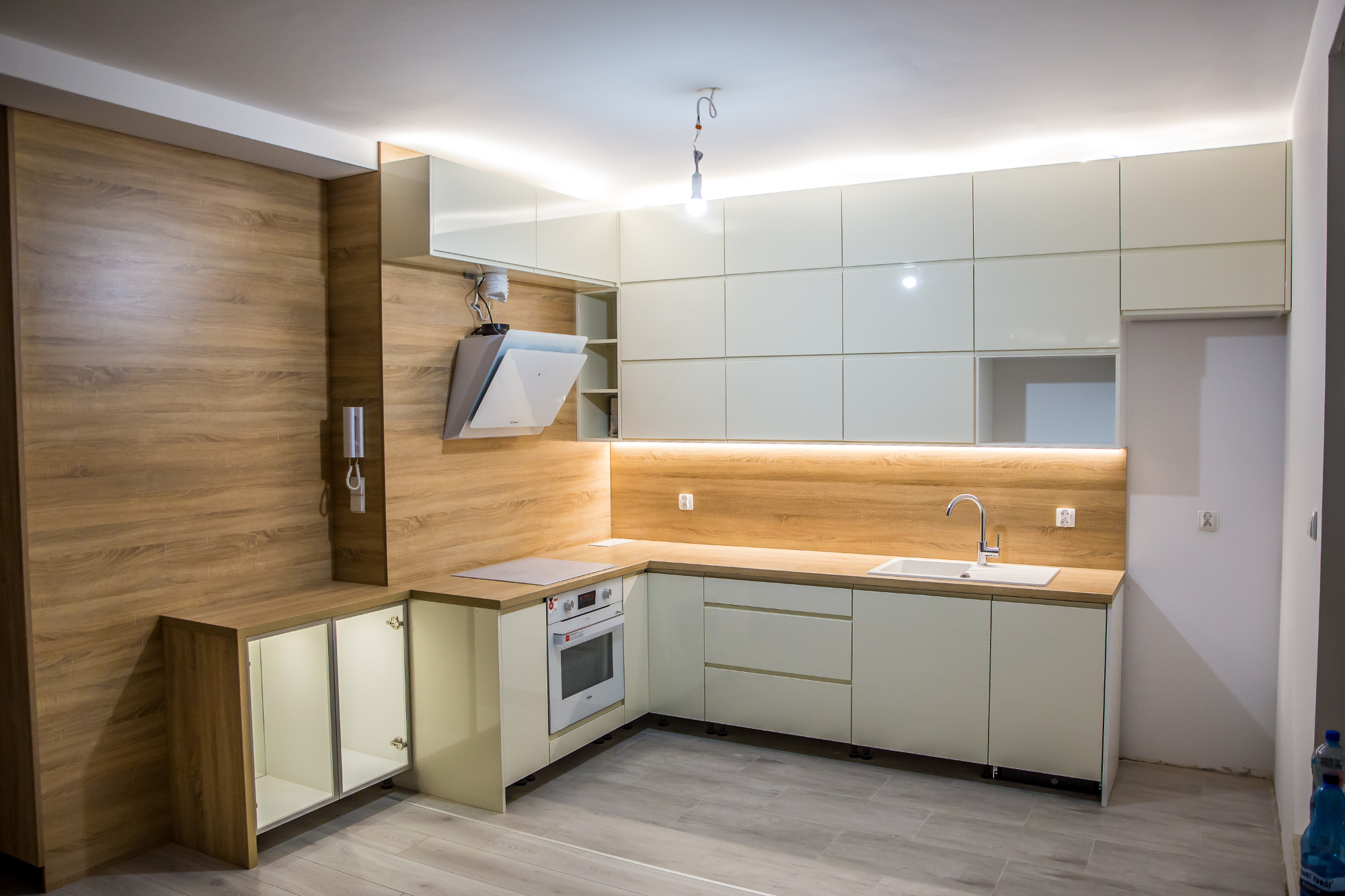 Nowoczesna kuchnia z białymi lakierowanymi frontami, drewnianym blatem i oświetleniem LED pod szafkami wiszącymi, widok na narożną zabudowę z piekarnikiem i okapem, bez dekoracji i...