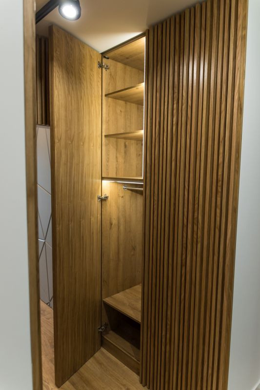 Otwarta szafa z drewnianymi lamelami na froncie, oświetlona od wewnątrz, z widocznymi półkami i drążkiem na ubrania.