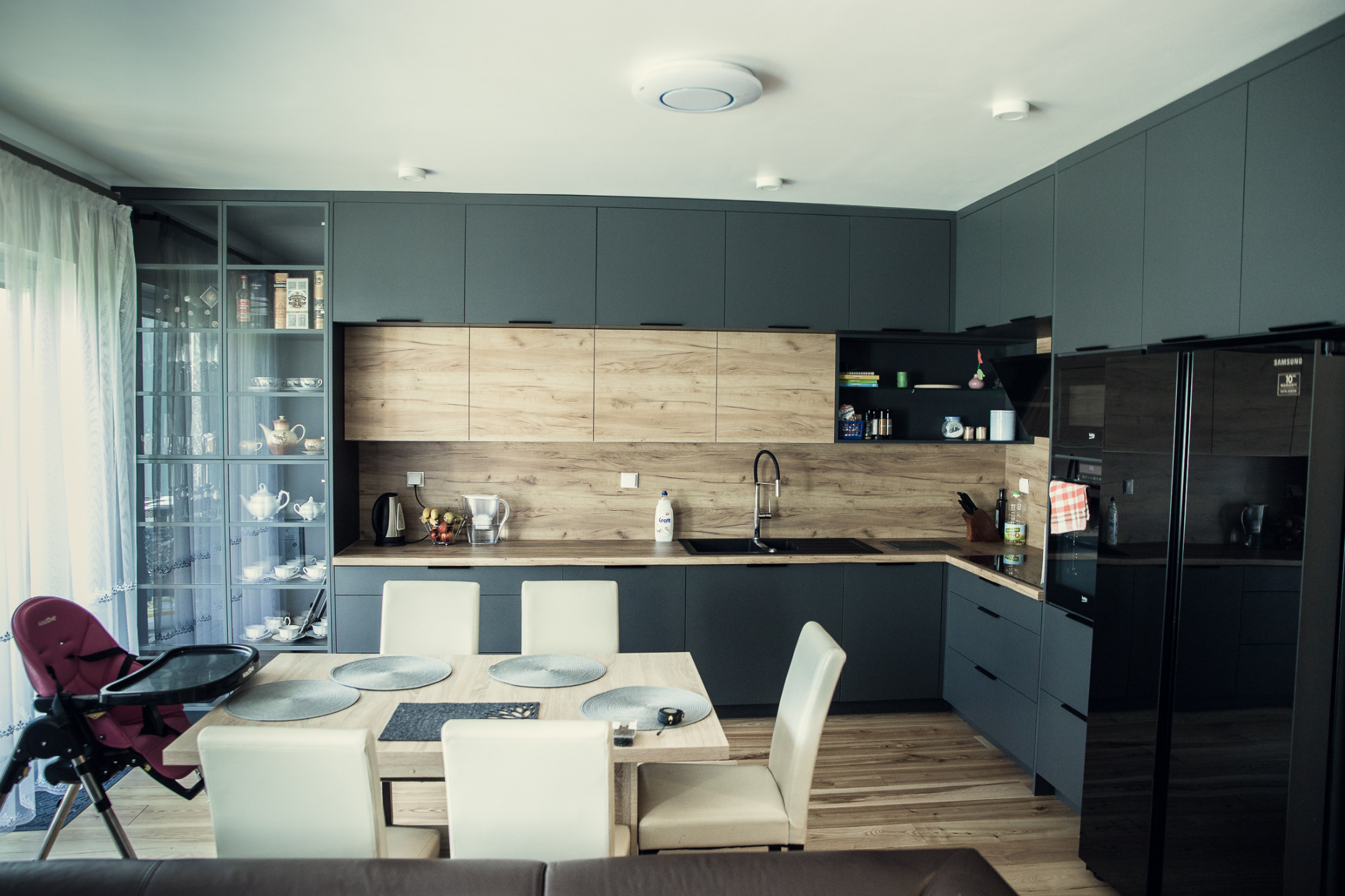 Nowoczesna kuchnia z szarymi szafkami i drewnianym blatem, szklana witryna z porcelaną, stół z białymi krzesłami i krzesełko dziecięce z boku.