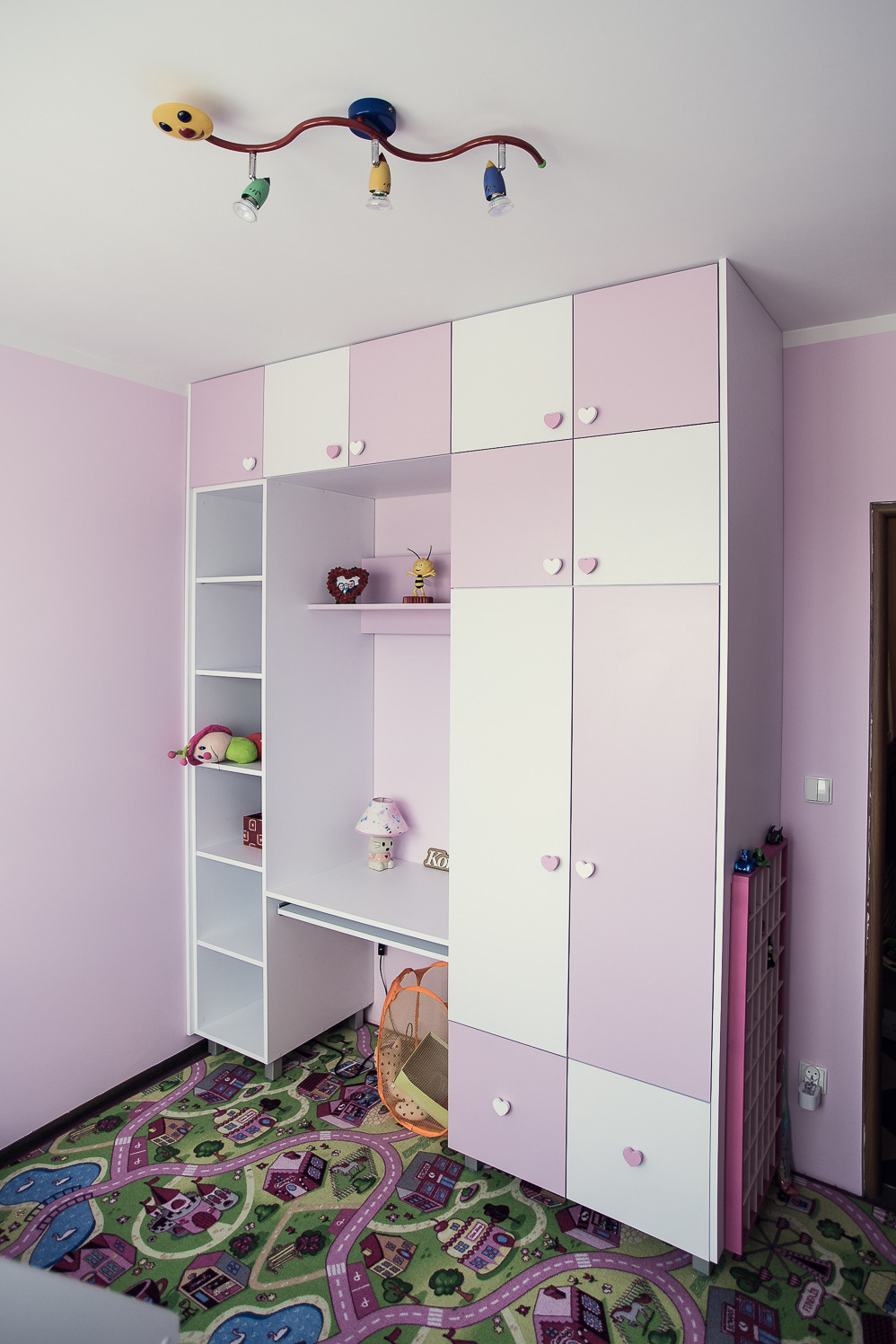 Różowo-biała zabudowa meblowa z szafami i biurkiem w pokoju dziecięcym, z uchwytami w kształcie serc, oświetlona lampą sufitową w kształcie uśmiechniętej buzi.