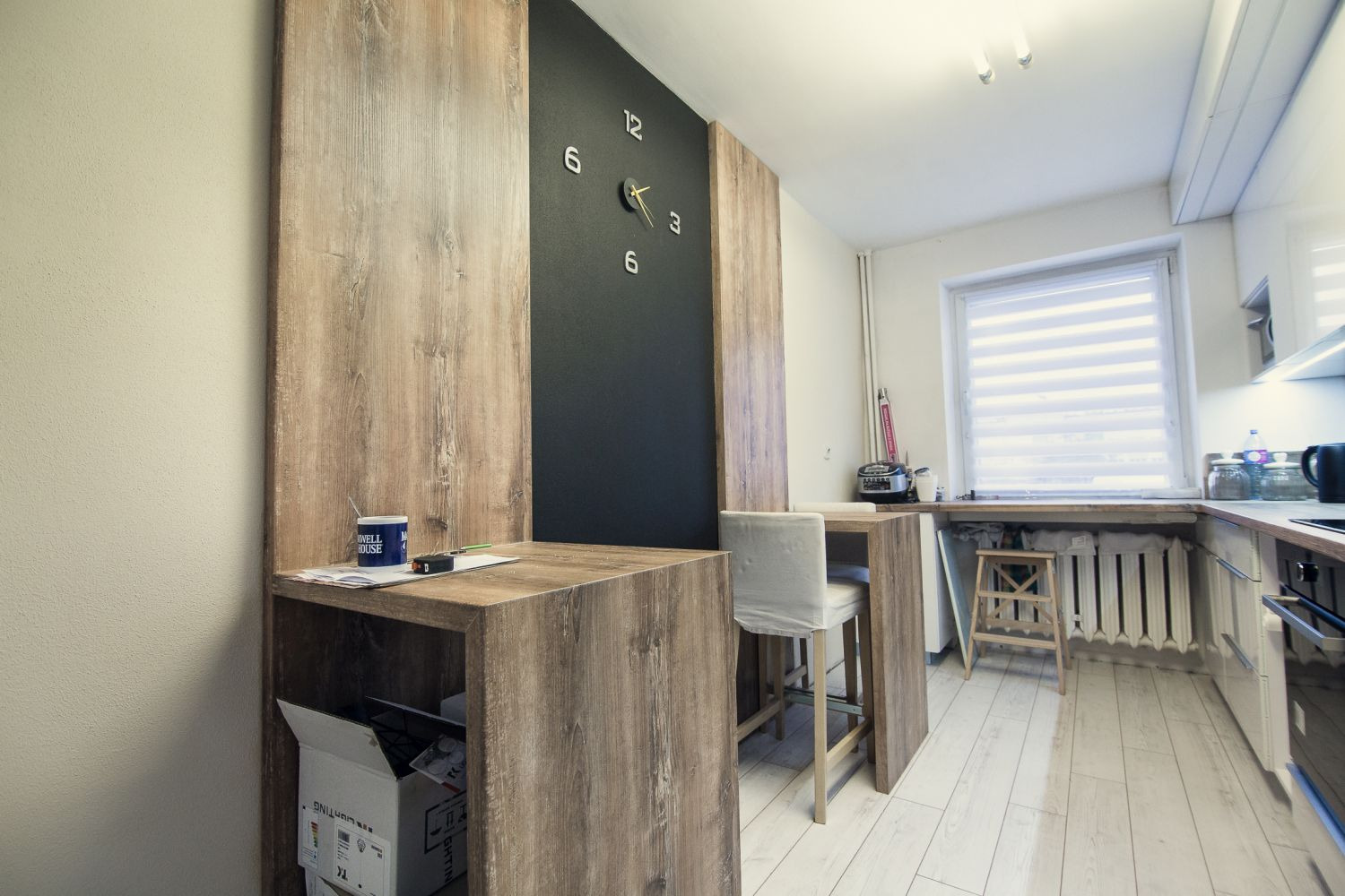 Nowoczesna kuchnia z drewnianym blatem barowym, czarną ścianą z zegarem i białymi krzesłami barowymi, oświetlona naturalnym światłem wpadającym przez okno z żaluzjami.