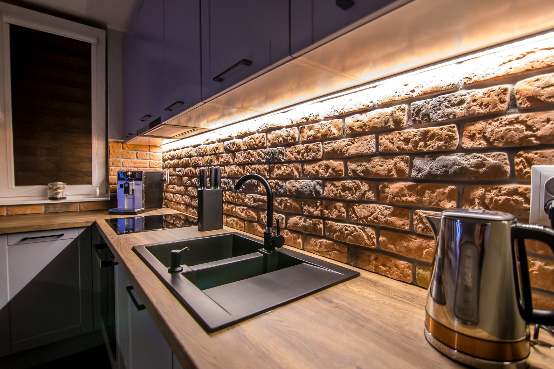 Nowoczesna kuchnia z czarnym, dwukomorowym zlewem, baterią i dozownikiem, drewnianym blatem, ścianą z cegły i oświetleniem LED podszafkowym.