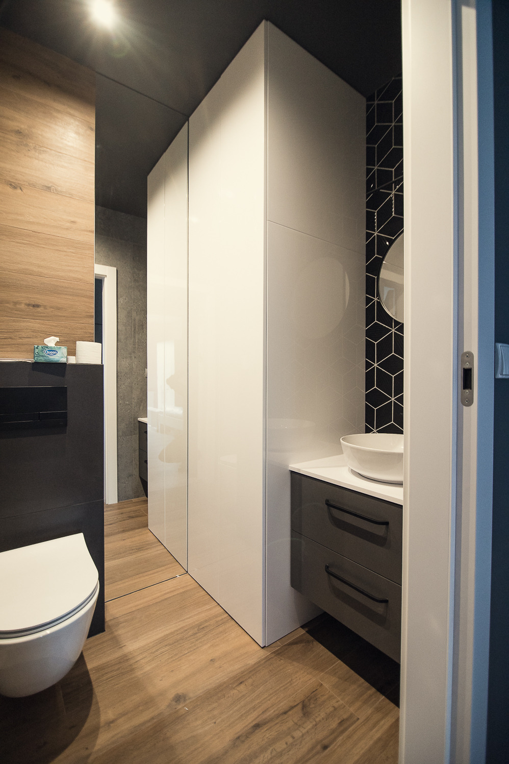 Nowoczesna łazienka z drewnopodobną podłogą, białą zabudową meblową, umywalką nablatową i czarno-białą mozaiką na ścianie