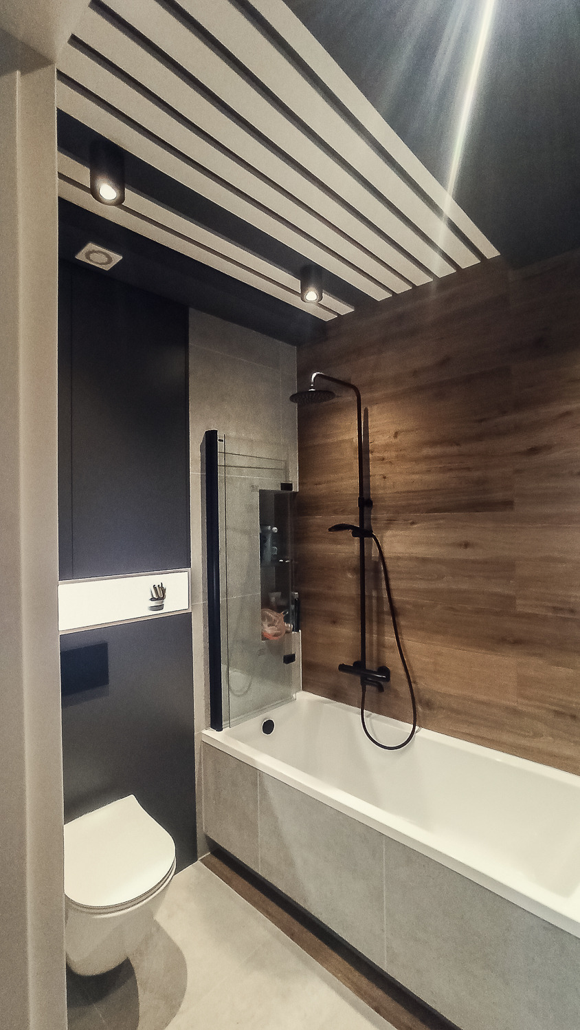 Nowoczesna łazienka z wanną, czarną armaturą i drewnopodobnymi panelami ściennymi, oświetlenie punktowe na suficie z dekoracyjnymi listwami.