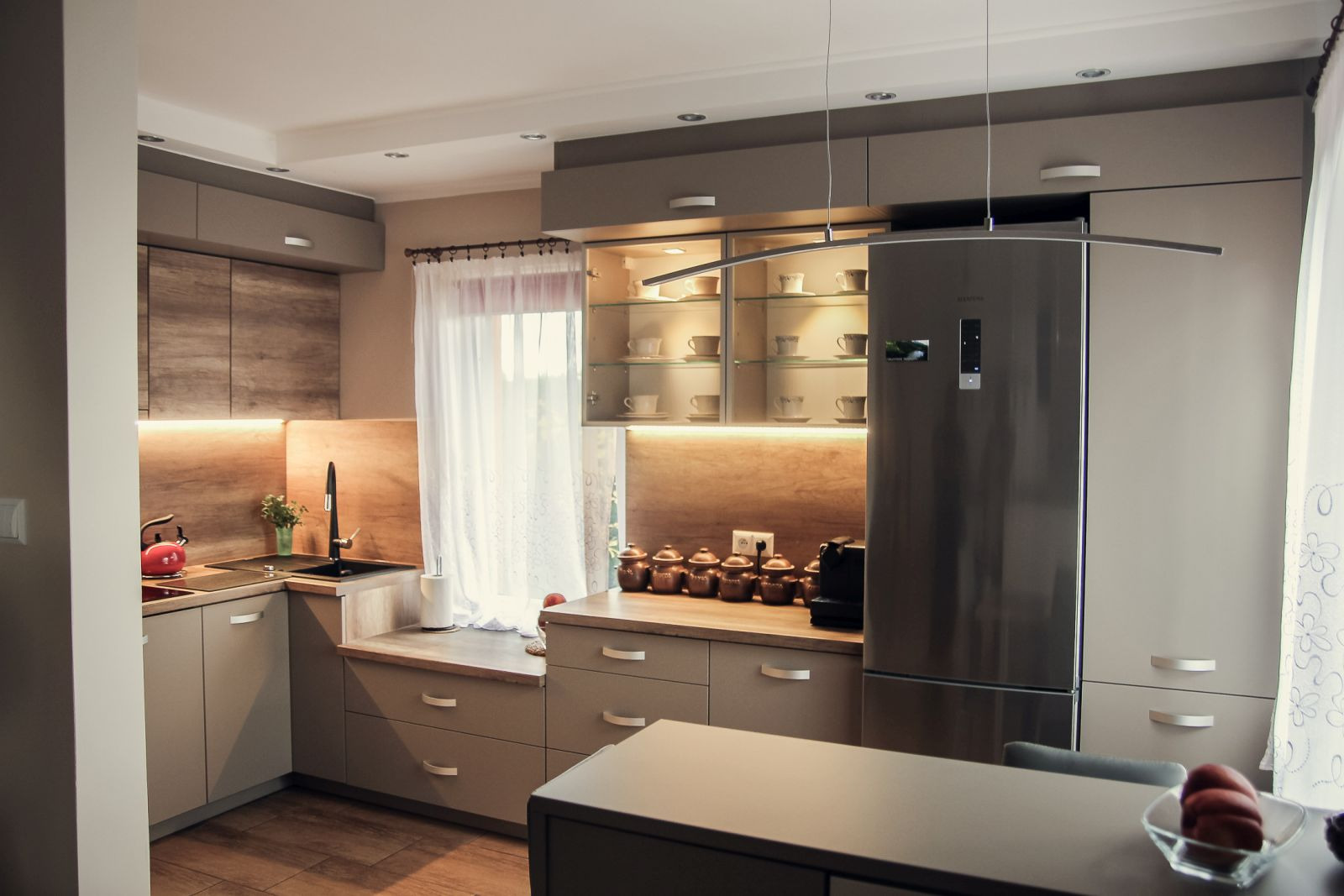 Nowoczesna kuchnia w odcieniach beżu i drewna z oświetleniem LED pod szafkami, szklana witryna z filiżankami i srebrna lodówka.