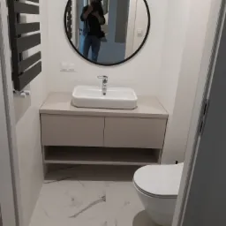 Nowoczesna łazienka z beżową szafką pod umywalką, okrągłym lustrem z odbiciem osoby robiącej zdjęcie i grzejnikiem drabinkowym.