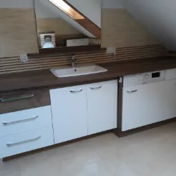 Zabudowa łazienkowa pod skosem z blatem imitującym drewno, białymi frontami szafek i wbudowaną pralką, odbicie okna dachowego w lustrze.