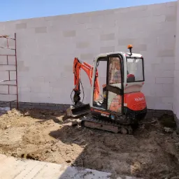 Pomarańczowa mini koparka Kubota KX41-3V w trakcie wyrównywania terenu na placu budowy, widoczne białe ściany z bloczków i metalowe rusztowanie.