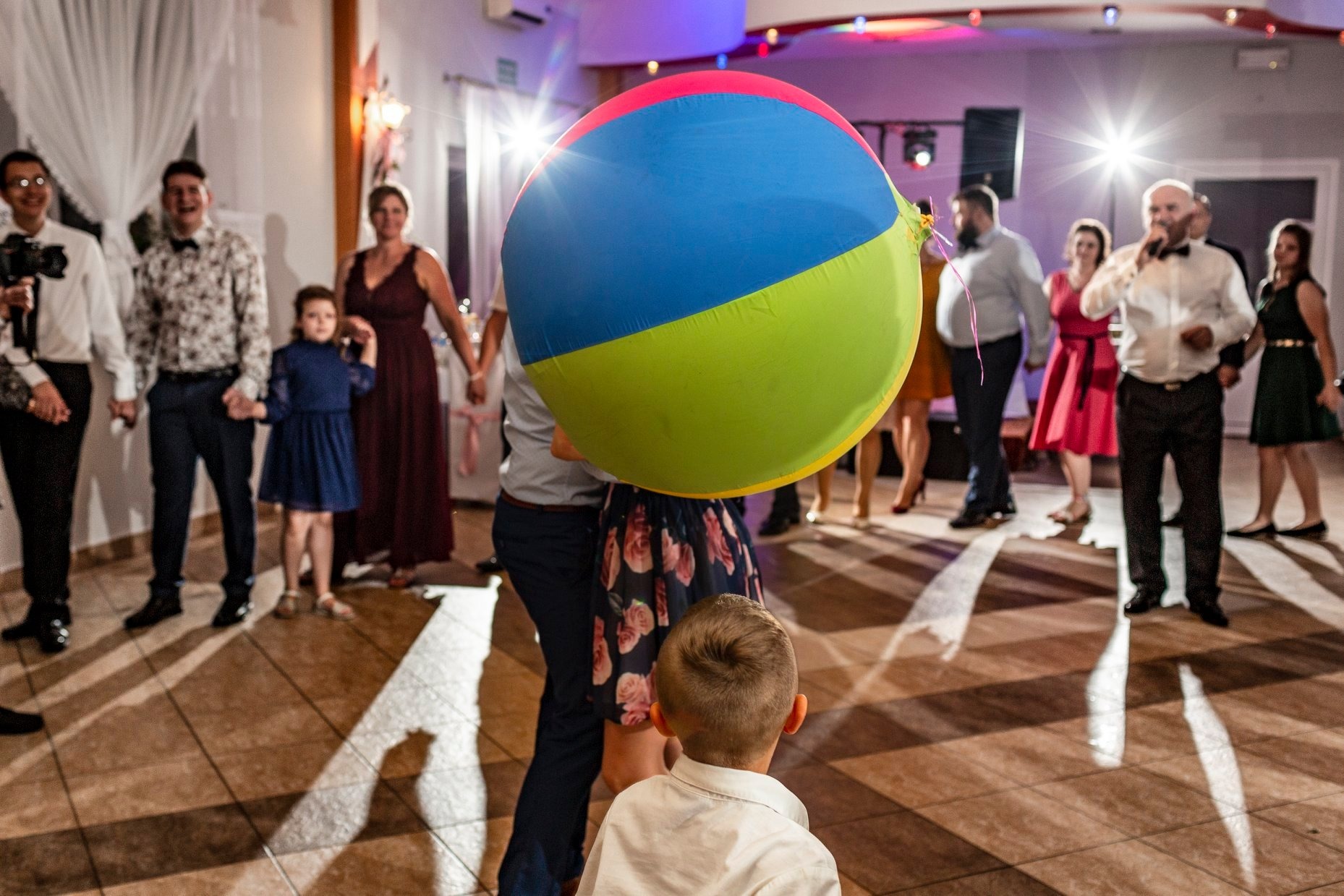 Duża, kolorowa piłka plażowa zasłaniająca parę podczas imprezy z udziałem dzieci, w tle widoczni goście i konferansjer.