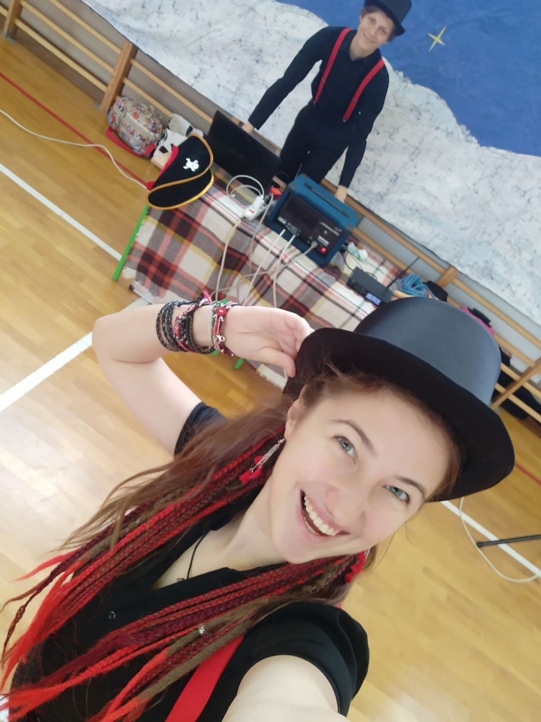 Uśmiechnięta kobieta w czarnym cylindrze i z czerwonymi warkoczykami pozuje na tle sceny z rekwizytami, w tle widoczna druga osoba również w cylindrze i szelkach.