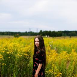 Natalia Mencfeld - Młoda kobieta w kwiecistej sukience pozuje na tle pola żółtych kwiatów, drzewa w oddali pod pochmurnym niebem.